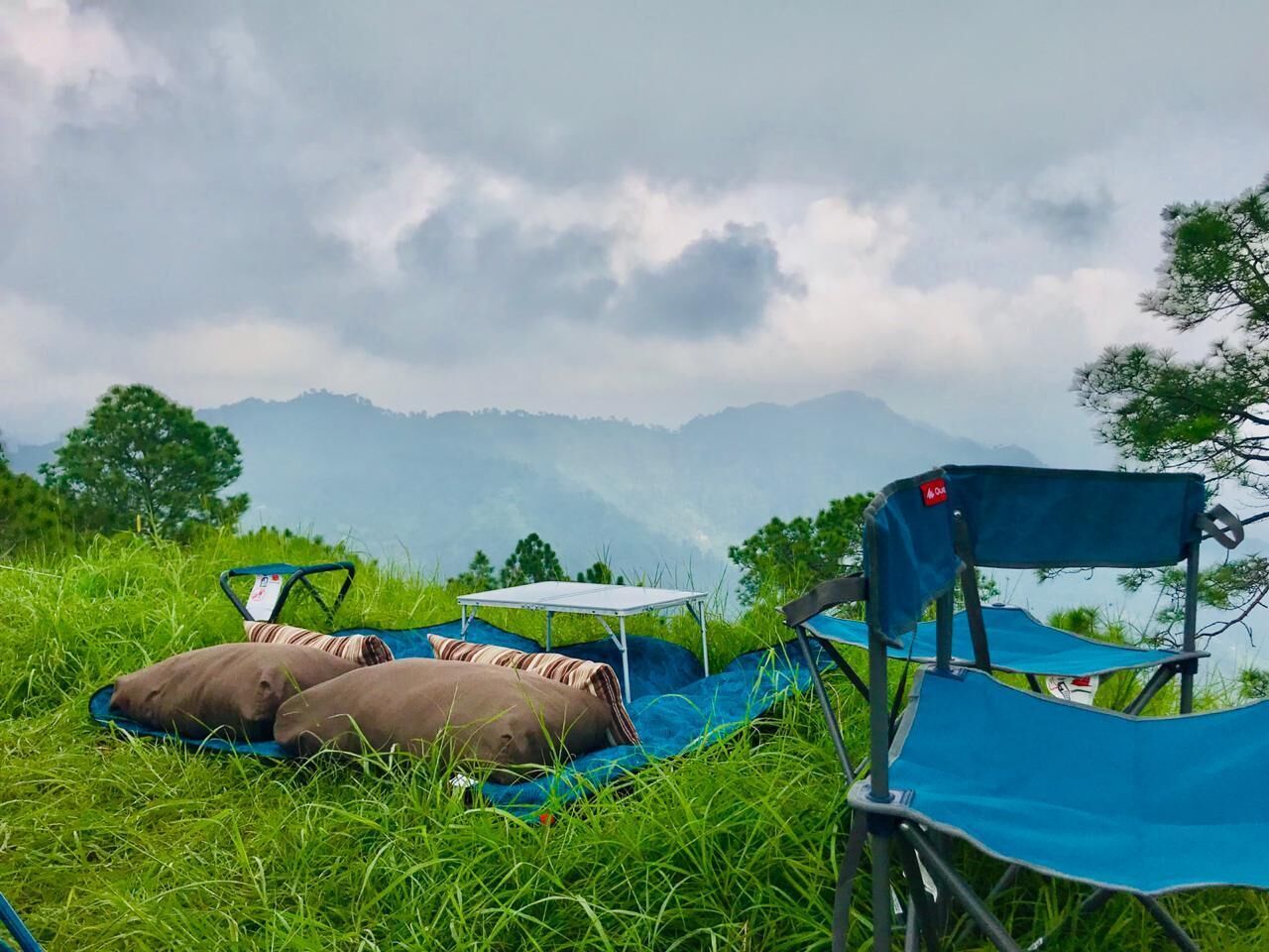 Un campamento con una tienda azul, una silla de camping y una mesa con cojín.