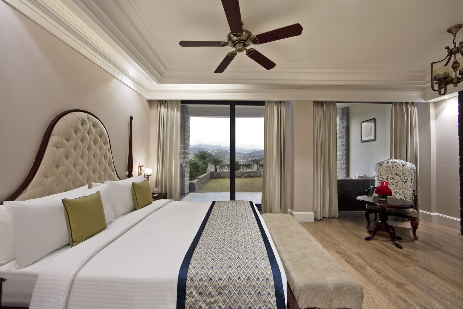 A spacious bedroom with a large bed, a ceiling fan, a view through the glass door, and a small table with a chair.