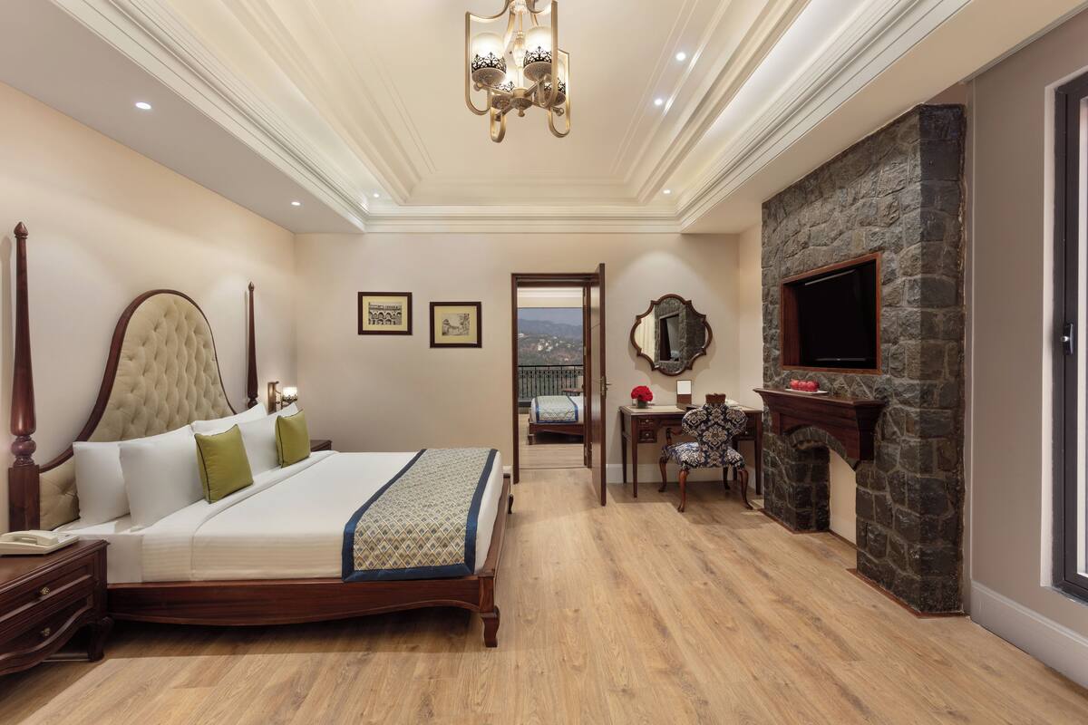 A bedroom with a large bed, a stone fireplace, a desk with a chair, and a view through an open door.