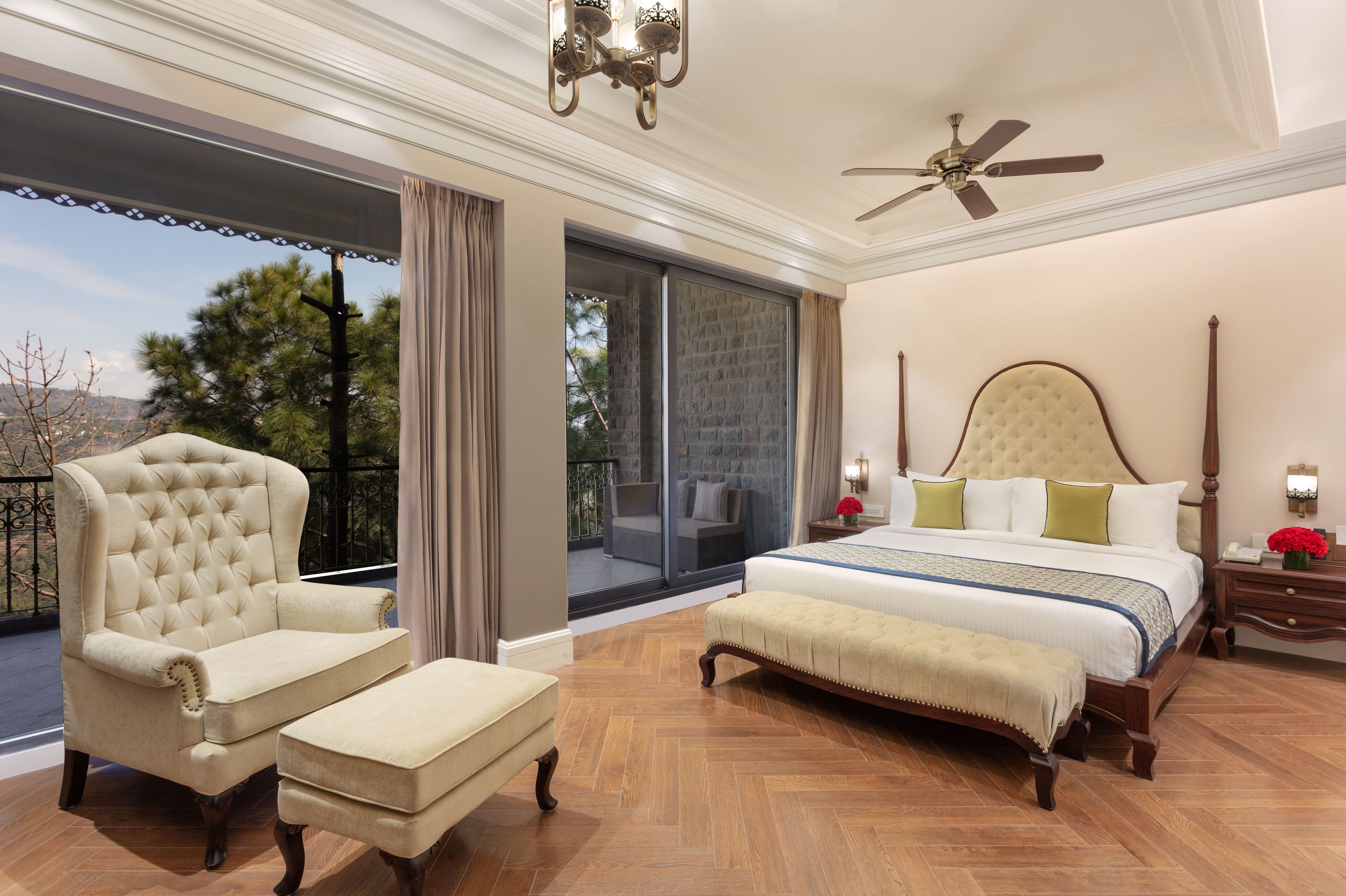 A spacious bedroom with a large bed, a sitting area with a chair and ottoman, a balcony with a view, and a ceiling fan.