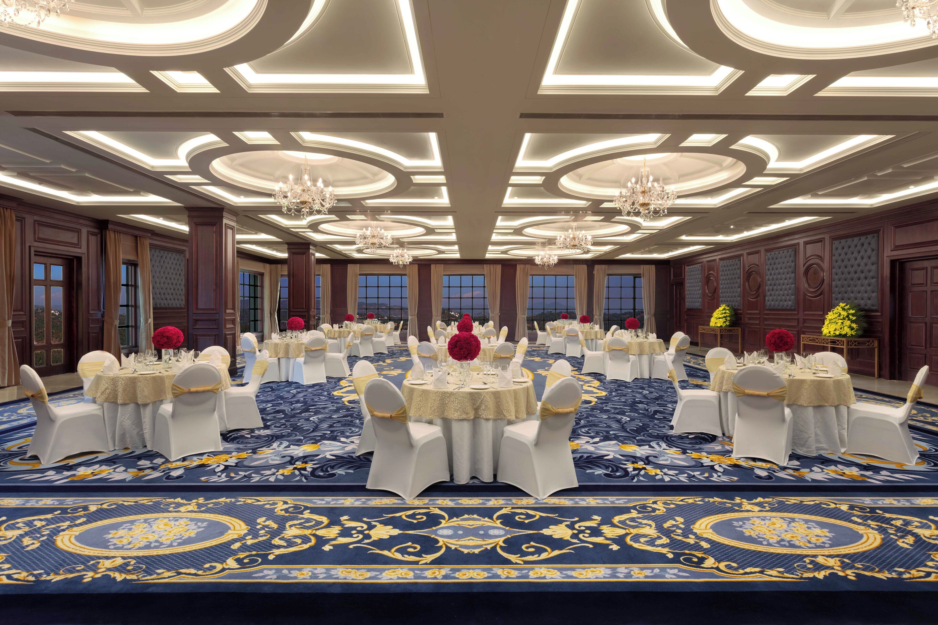 Un salón de banquetes con mesas redondas, sillas blancas, candelabros y una alfombra azul con detalles dorados.