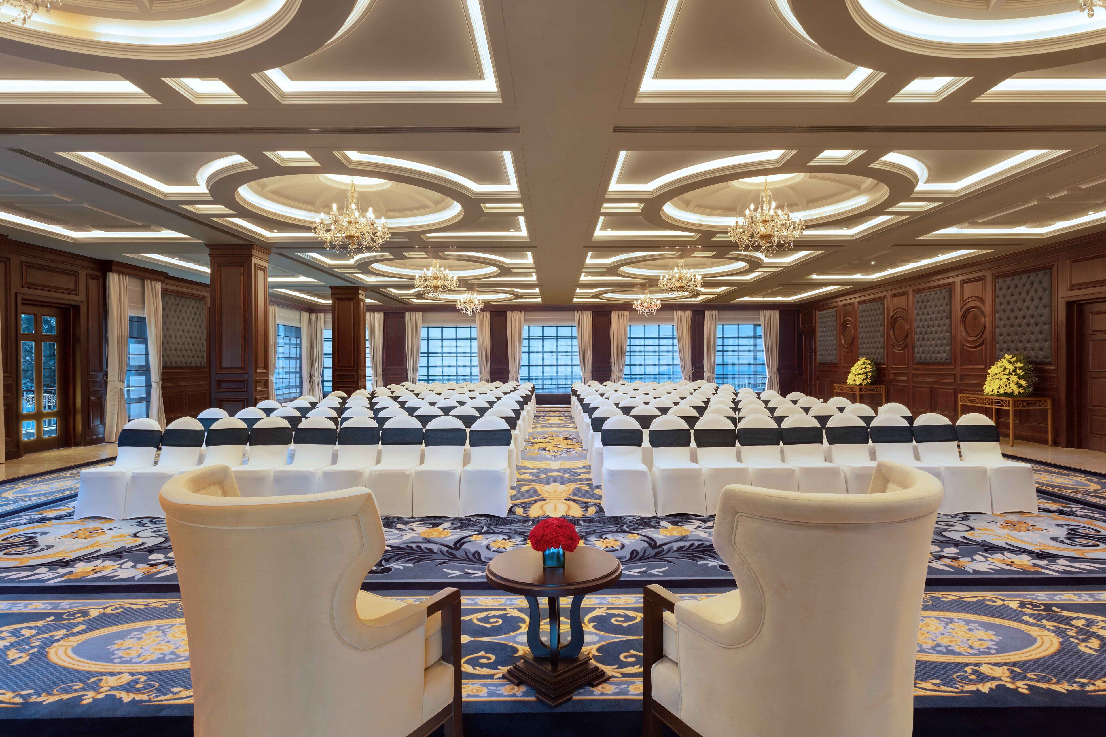 Une salle de conférence avec des rangées de chaises blanches, une table centrale ornée d’un vase de fleurs et des lustres au plafond.