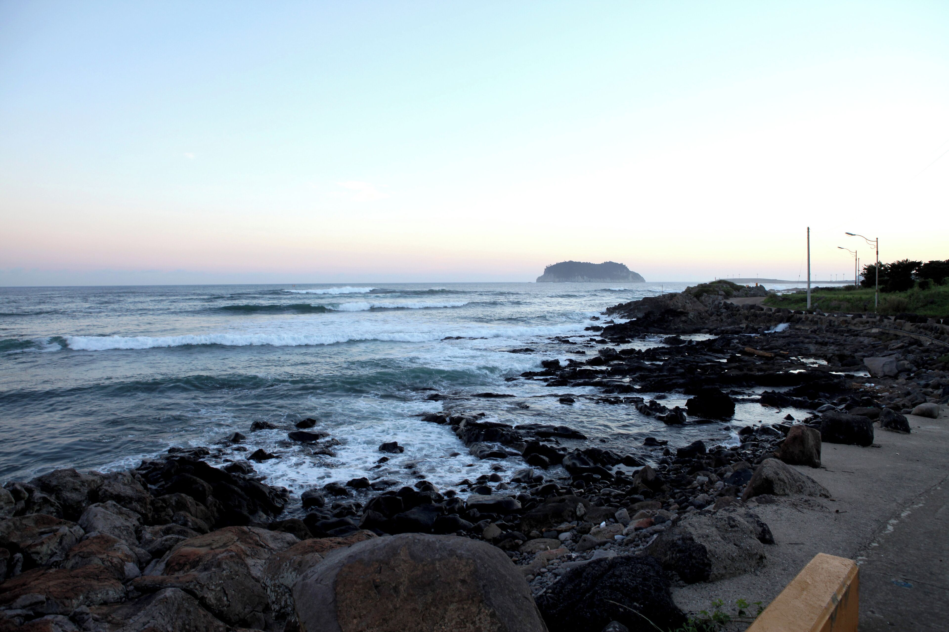 Una costa rocosa con olas rompiendo sobre la playa, una isla distante y un camino pavimentado a lo largo de la costa.