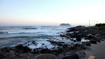 Una costa rocosa con olas rompiendo sobre la playa, una isla distante y un camino pavimentado a lo largo de la costa.