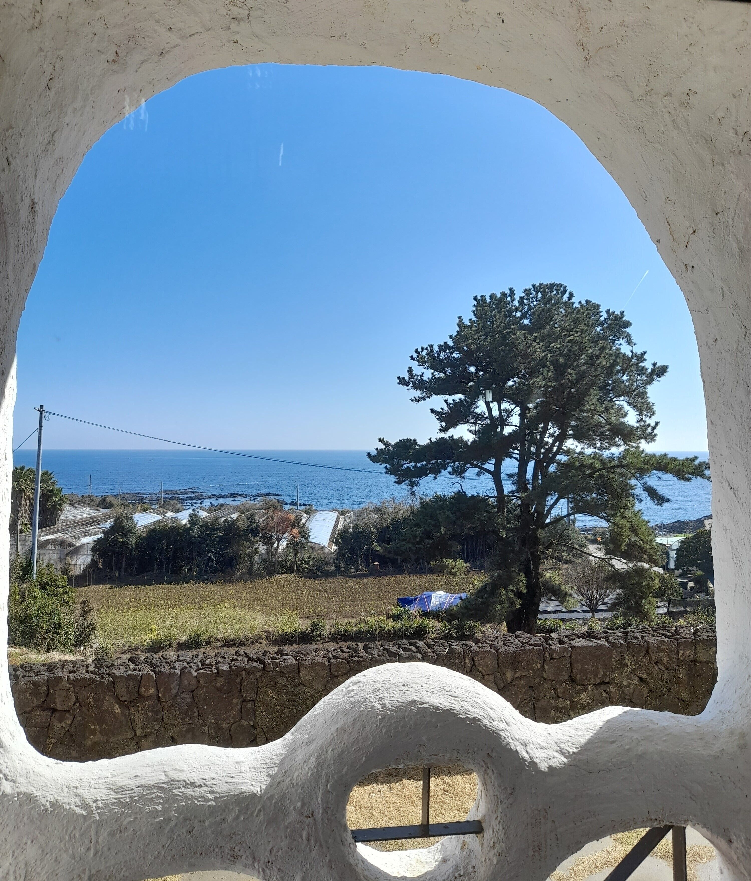 A view through a window with a stone wall and a tree, overlooking a coastal area with fields and a clear sky.