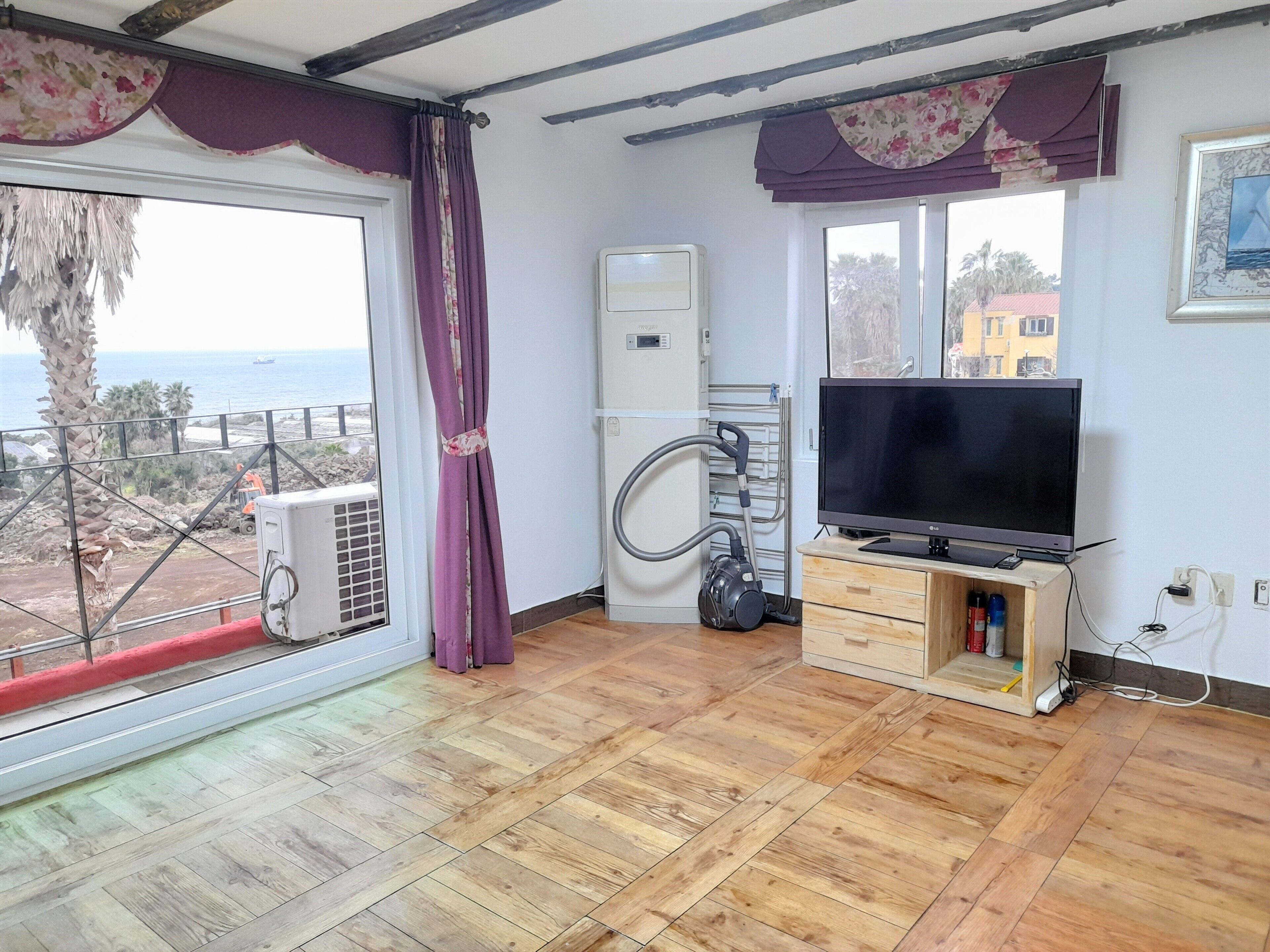 A room with a wooden floor, a TV on a wooden stand, a window with a view of the sea, and a ceiling fan.