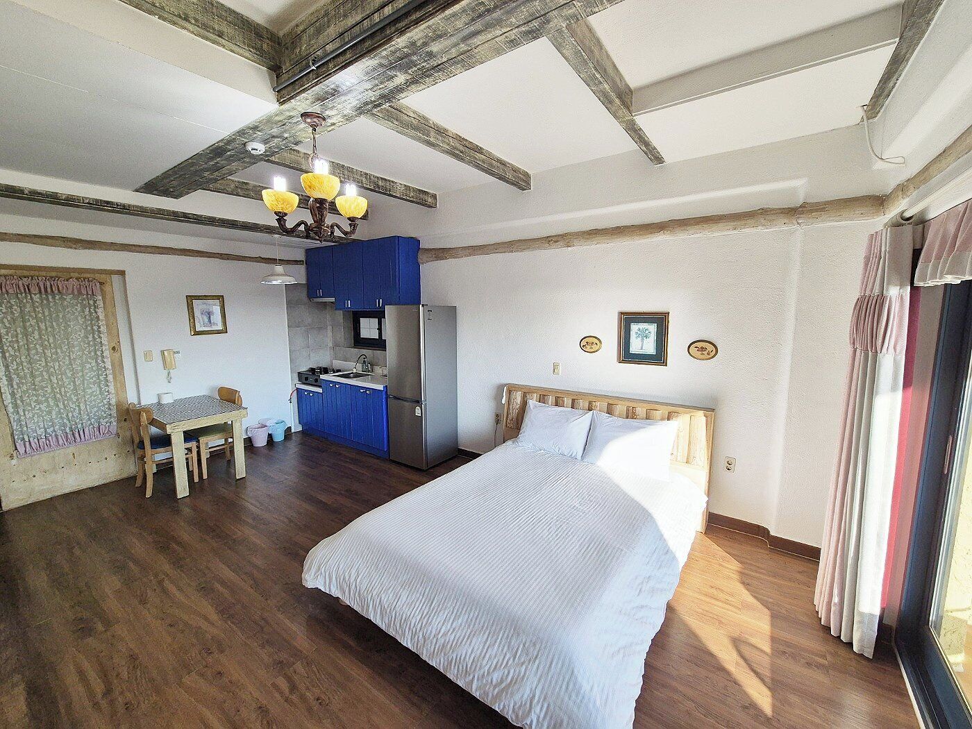 A bedroom with a bed, a wooden headboard, a desk with a chair, a kitchenette with blue cabinets, and a ceiling with exposed beams.