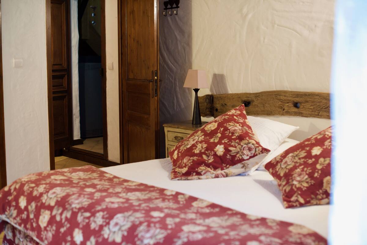 Une chambre à coucher avec une tête de lit en bois, un lit recouvert d’une literie à motifs floraux rouges, une table de chevet avec une lampe, et une porte entrouverte donnant sur une autre pièce.