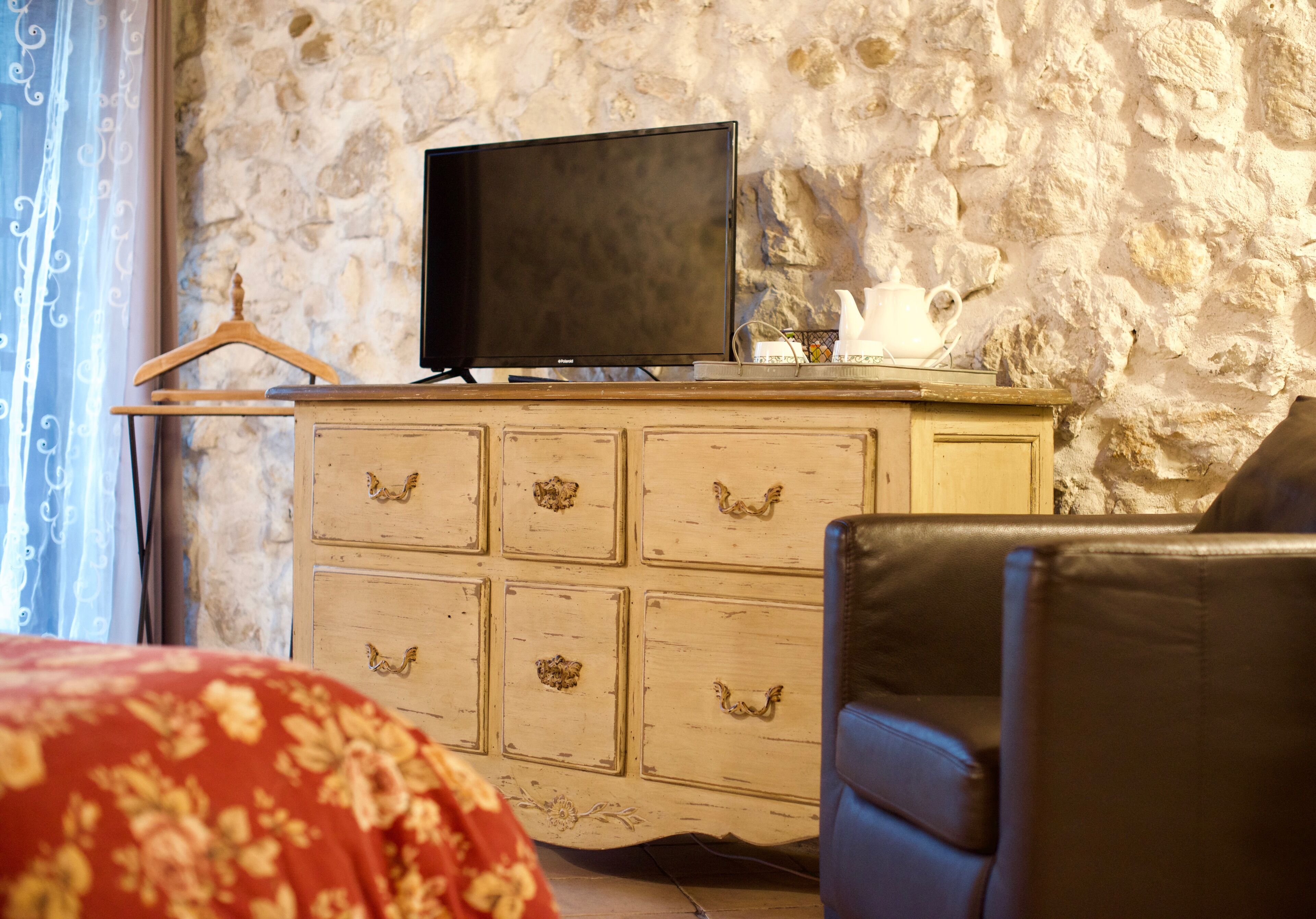 Une pièce avec un téléviseur posé sur une commode en bois, un fauteuil en cuir noir et une couvre-lit à motifs floraux rouges.