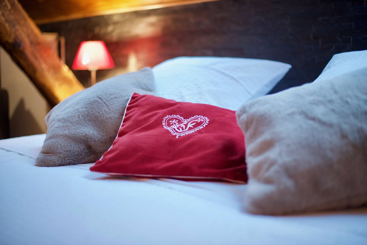 Un lit avec des oreillers blancs et rouges, un oreiller rouge orné d’un motif en forme de cœur, et une lampe rouge en arrière-plan.