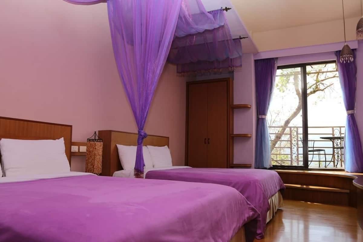 A room with two beds, a wooden headboard, a window with curtains, and a balcony with a table and chairs.