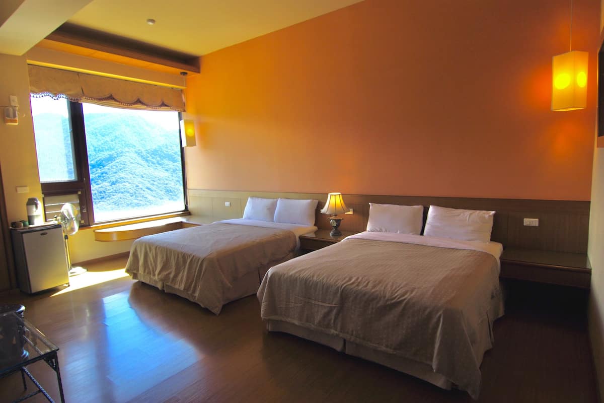 A hotel room with two beds, a desk, a lamp, and a window with a mountain view.
