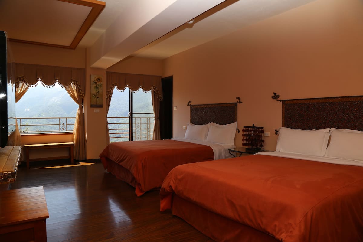 A hotel room with two beds, a wooden bench, a small table, and a view of the mountains through the window.