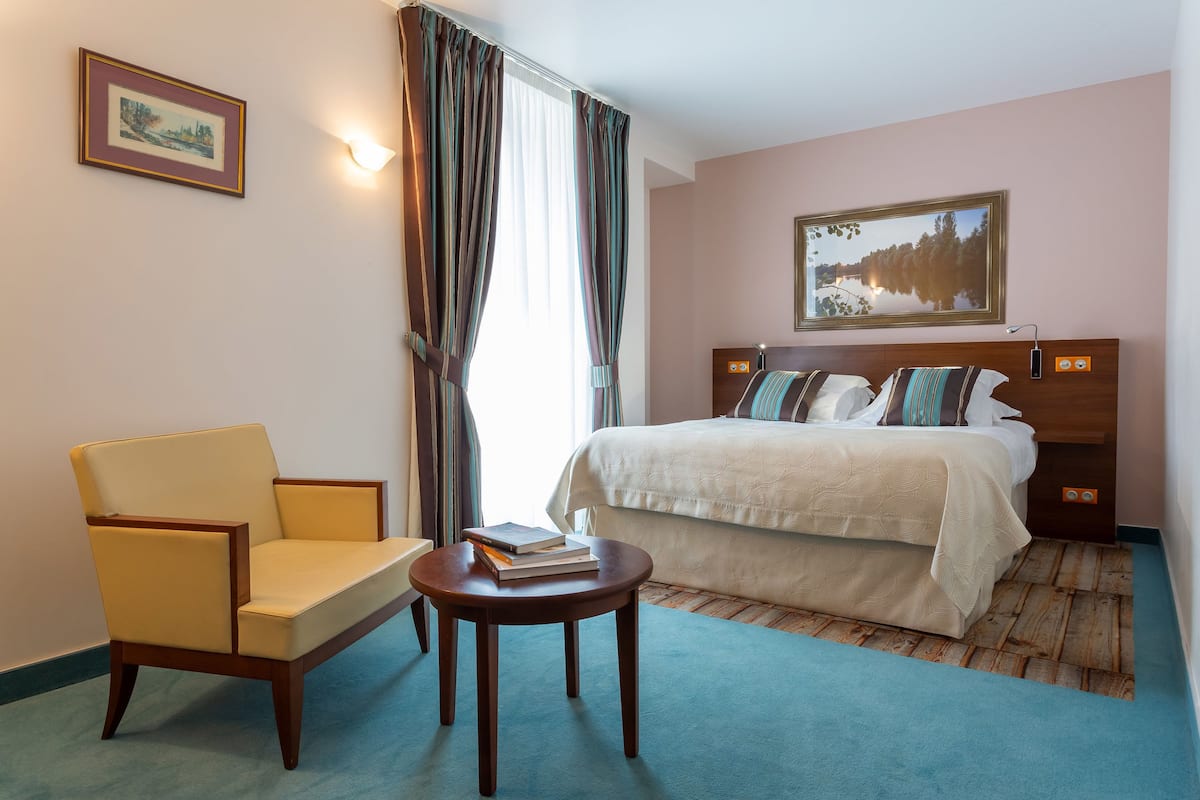 A hotel room with a bed, a chair, a small table, and a painting on the wall.