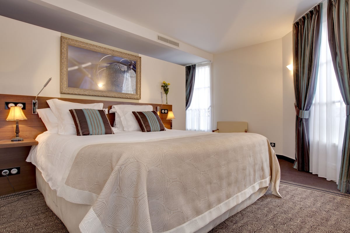 A hotel room with a large bed, a headboard with a mirror, a bedside table with a lamp, and a window with curtains.