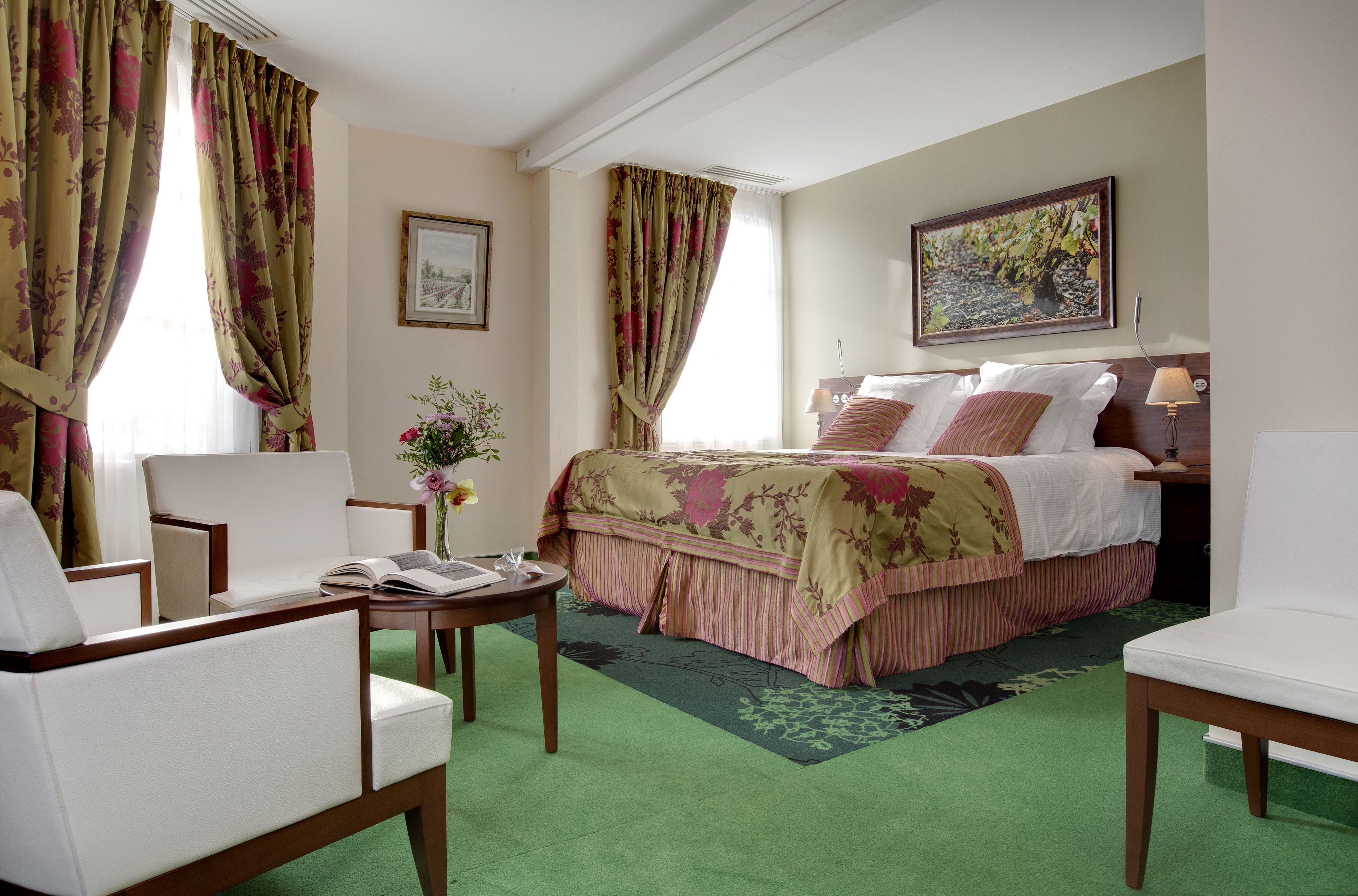 Hotelkamer met een bed, twee stoelen, een kleintje tafeltje, een vaas met bloemen en een schilderij aan de muur.