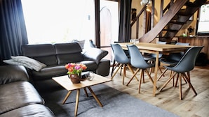 A modern living room with a black leather sofa, a wooden coffee table with a vase of flowers, and a dining area with wooden chairs and a table. - Ferienhaus Drebach (Drebach)