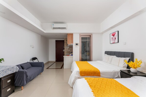 A modern hotel room with two beds, a sofa, a desk, and a bathroom visible through an open door.
