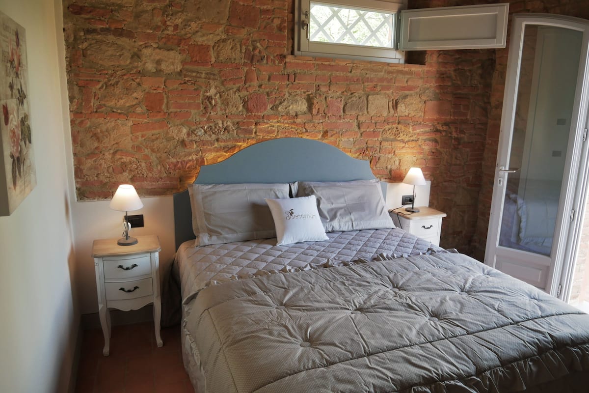 A bedroom with a brick wall, a bed with a quilted headboard, two bedside tables with lamps, and a wardrobe.