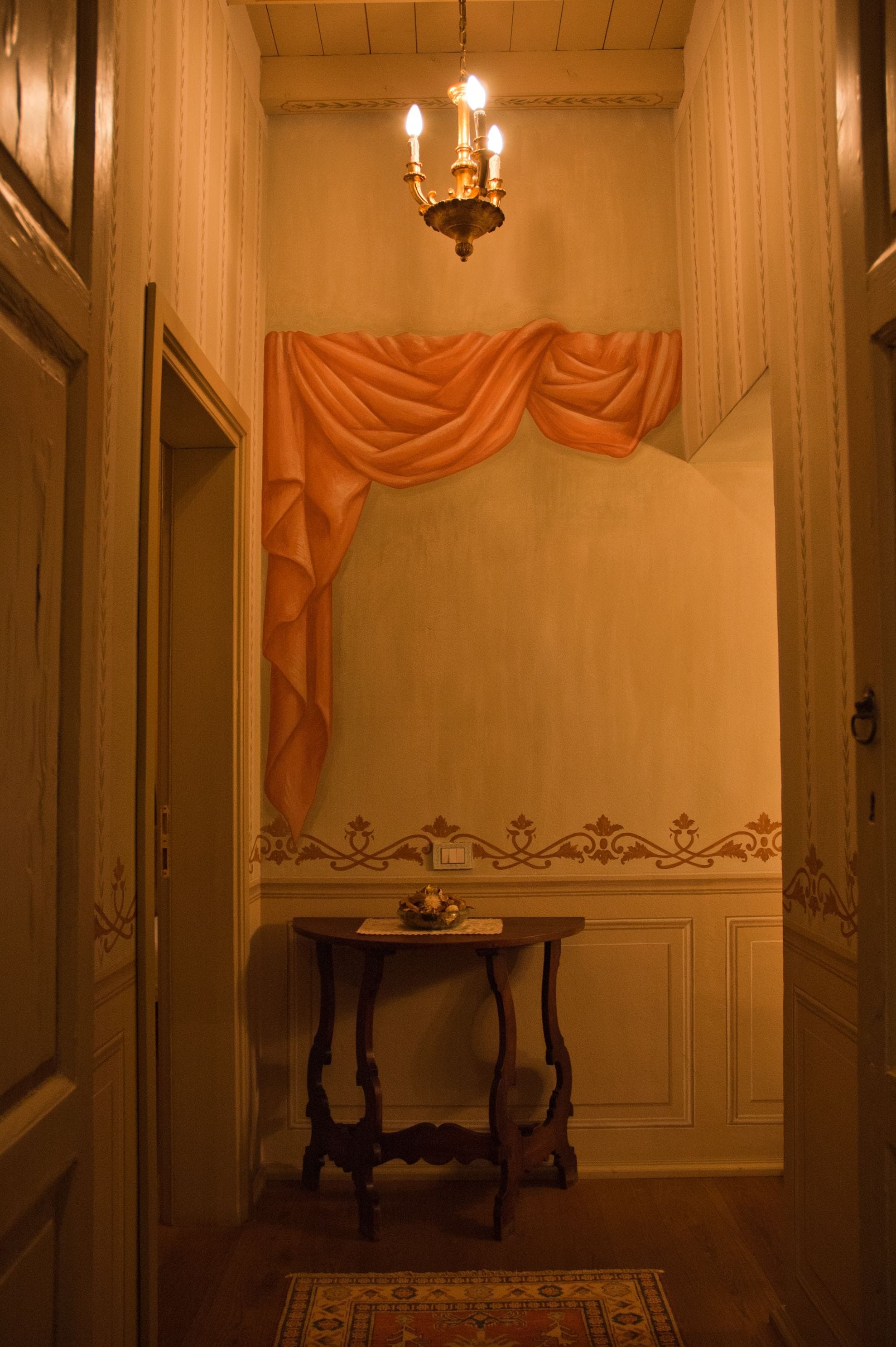 Un corridoio con un lampadario, una tenda drappeggiata, un tavolino con un oggetto decorativo e un tappeto a motivi.