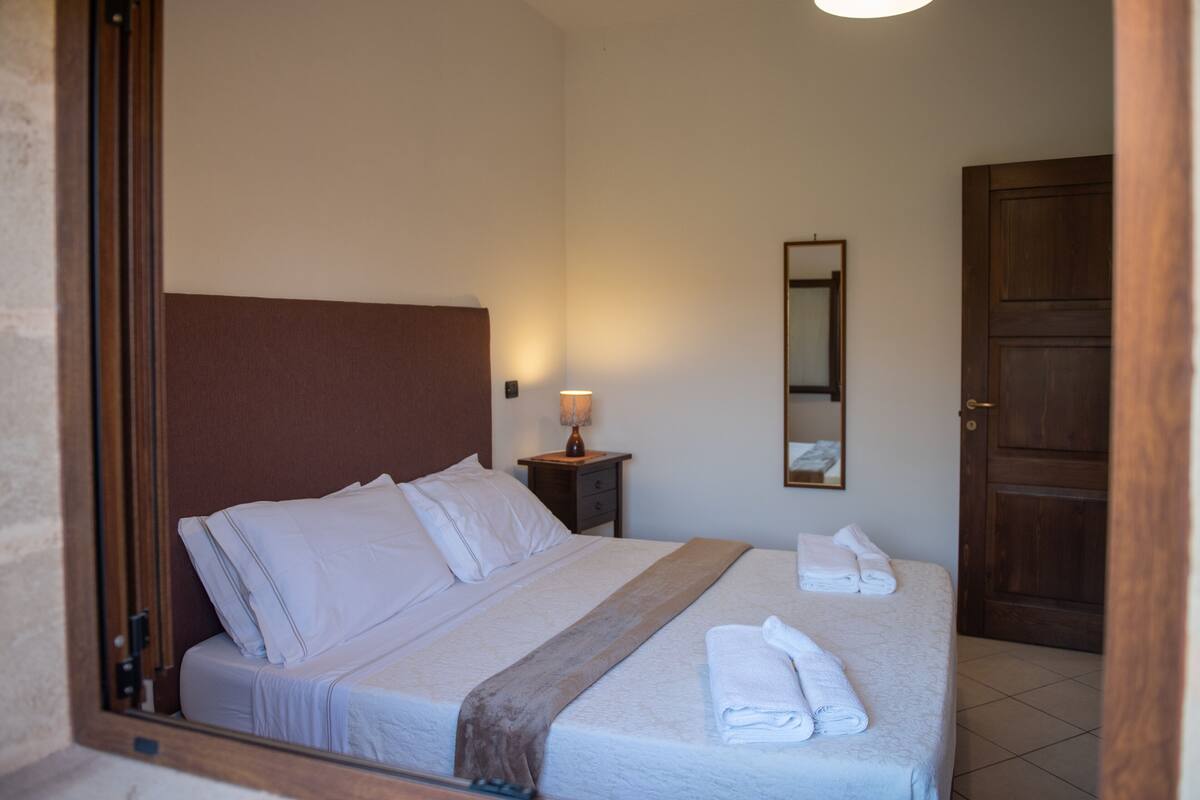 A neatly made bed with white linens and a brown headboard, a bedside table with a lamp, and a wooden door.