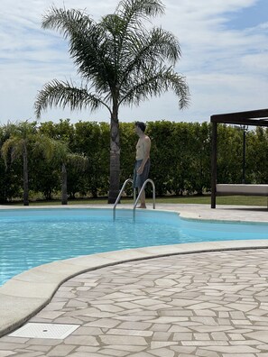 Un hombre de pie junto a una piscina, con una palmera y una pérgola al fondo.