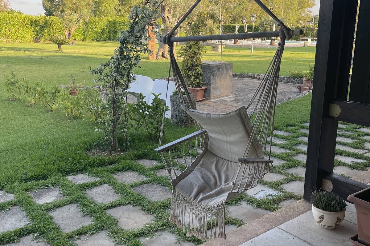 A hammock chair under a pergola with a view of a grassy park and trees.