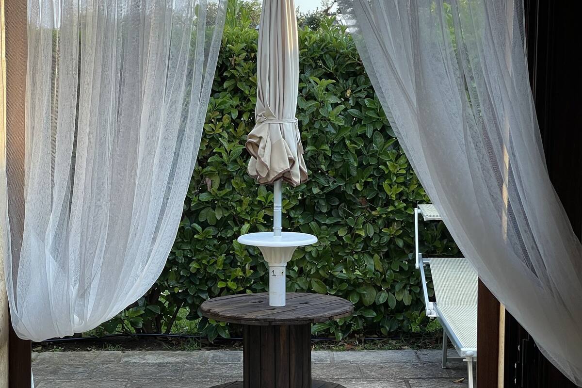 An outdoor patio with a round table and an umbrella, surrounded by greenery and a stone floor.