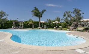 Una piscina con sillones de descanso y sombrillas, rodeada de árboles y una pérgola.