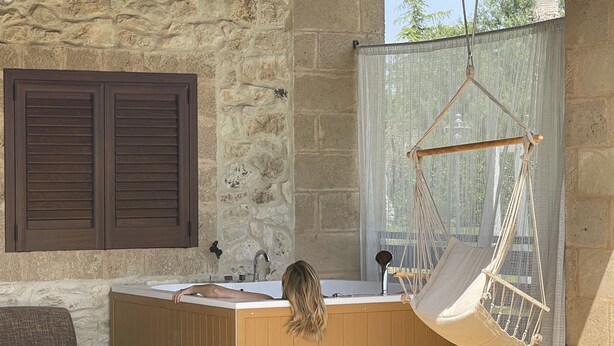 A stone-walled room with a wooden bathtub, a hanging hammock, and a window with curtains. - Masseria Giamarra (Carpignano Salentino)