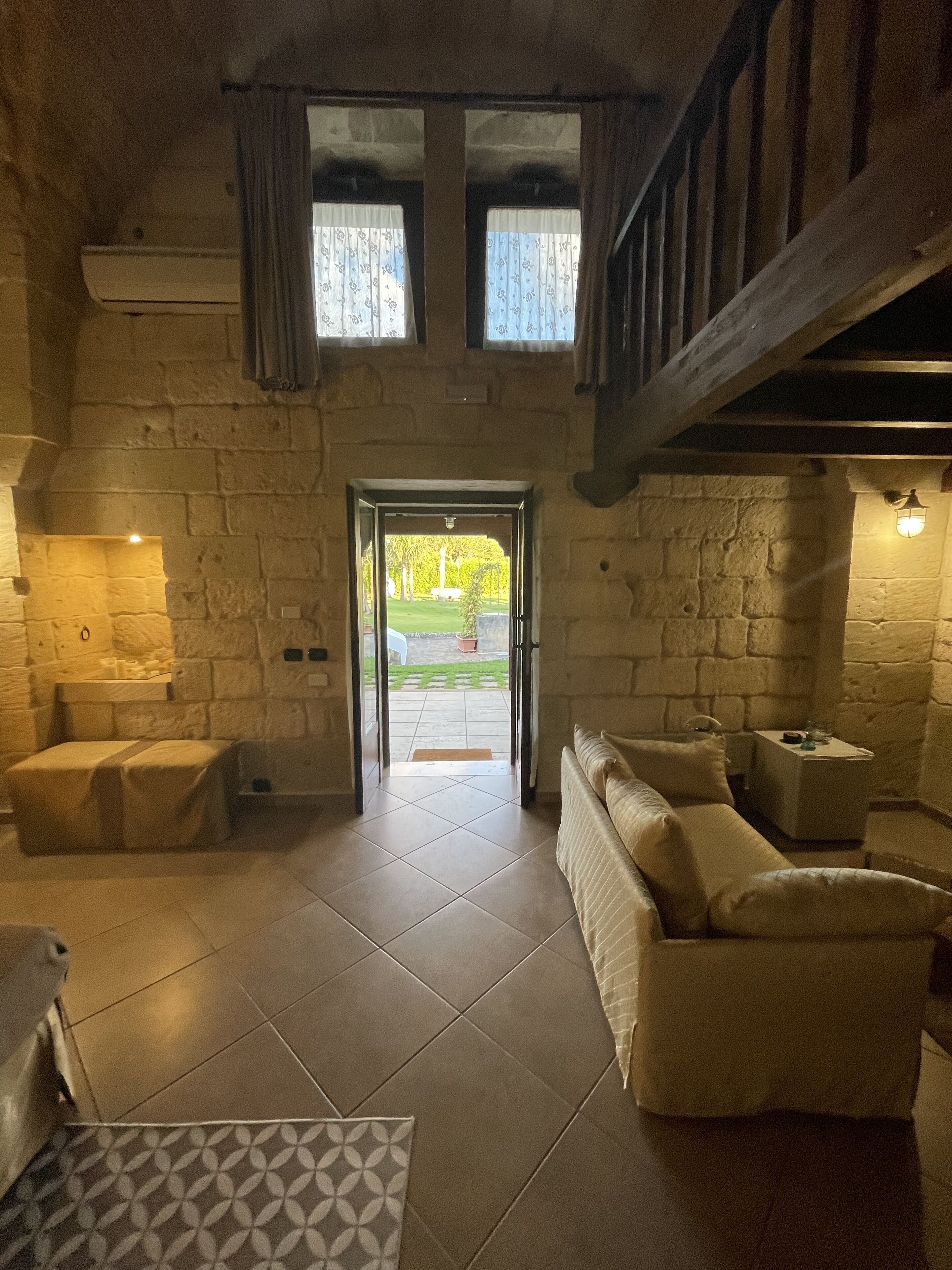 A rustic interior with stone walls, a wooden staircase, a sofa, and a tiled floor.