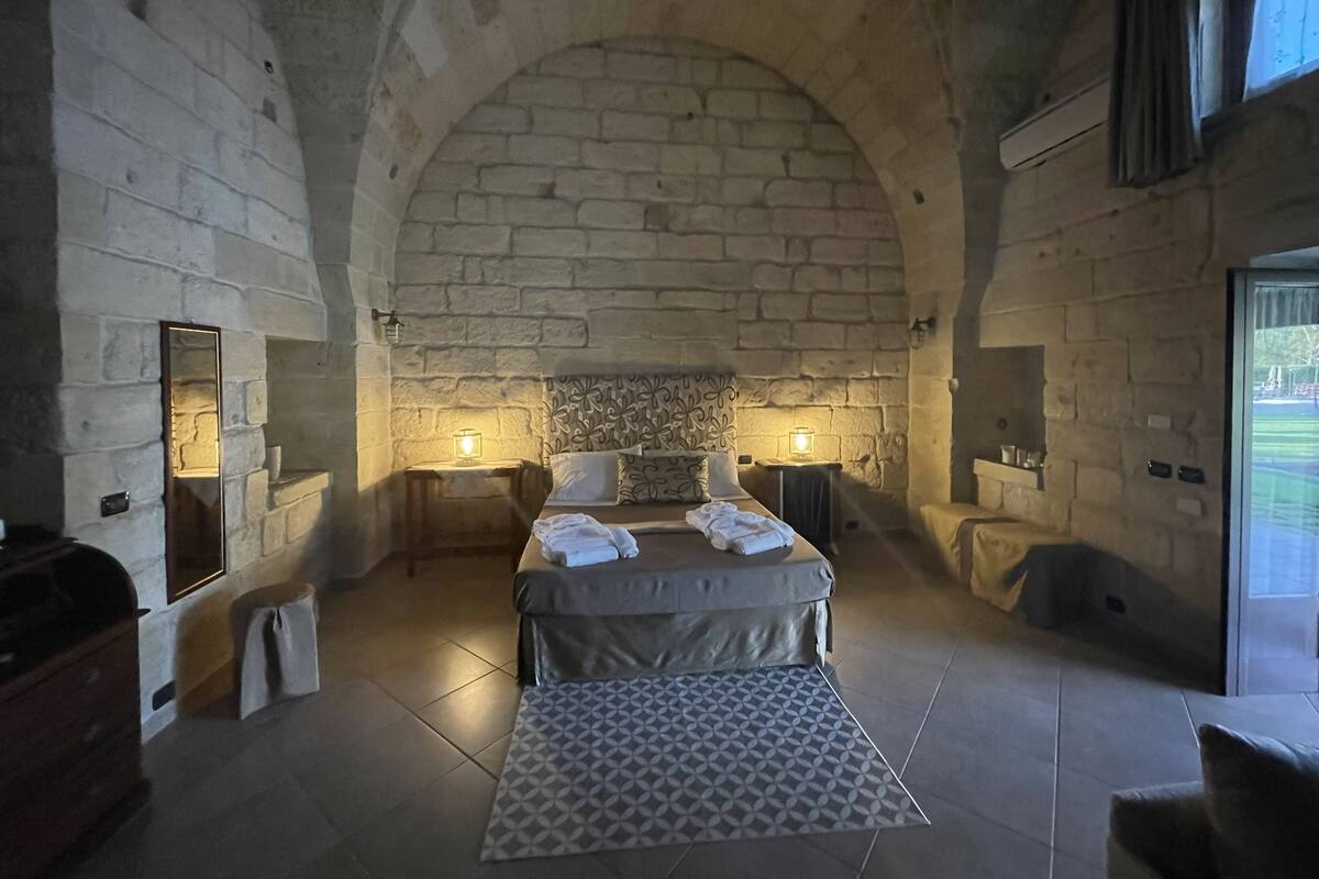 A bedroom with a stone wall, a bed with a patterned rug, two bedside tables with lamps, and a window with curtains.