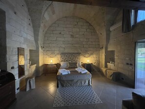 A bedroom with a stone wall, a bed with a patterned rug, two bedside tables with lamps, and a window with curtains. - Masseria Giamarra (Carpignano Salentino)