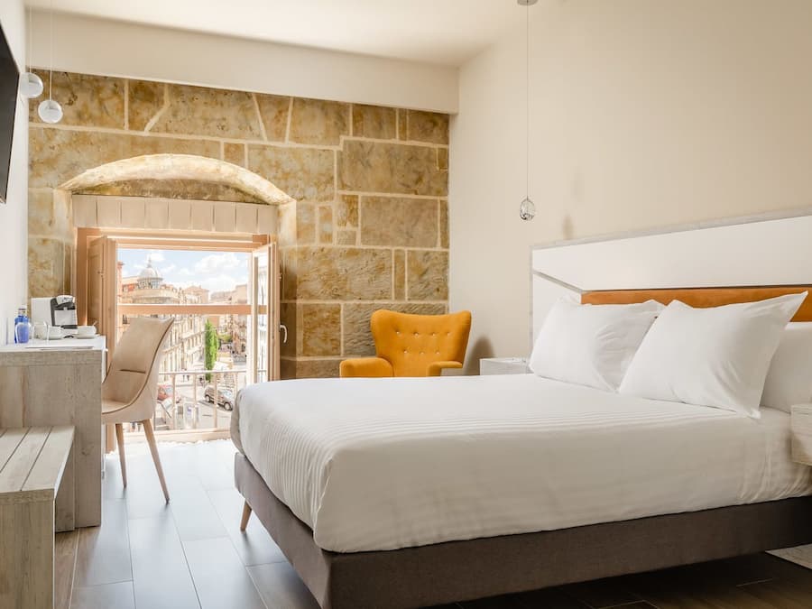 Habitación de hotel con una cama grande, un escritorio, una silla y vista a la ciudad a través de un arco abierto.