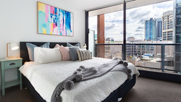Une chambre moderne avec un grand lit, un tableau abstrait aux couleurs vives et une vue sur la ville à travers de grandes fenêtres.