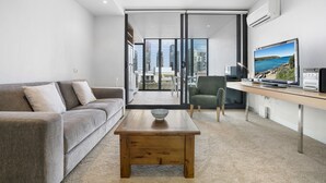 A modern living room with a grey sofa, a wooden coffee table, a TV, and a view of the city through large windows. - Leila, 1BDR Melbourne Apartment (Melbourne)
