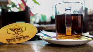 A glass of amber liquid on a white saucer, a wooden coaster with a logo, and a blurred background with greenery. - Quetzel Guest House (Pokhara)