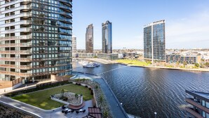 A high-rise building with a curved facade, a river with a boat dock, and a landscaped area with seating. - Sabrina, 2BDR Docklands Apartment (Docklands)