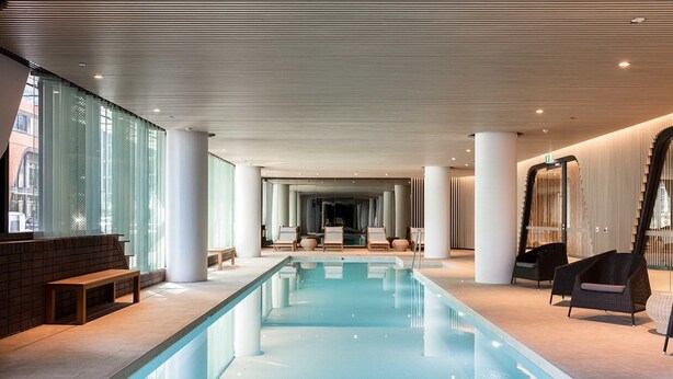 A modern indoor swimming pool with clear glass walls, lounge chairs, and a central seating area. - Sabrina, 2BDR Docklands Apartment (Docklands)