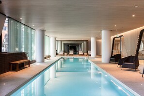 A modern indoor swimming pool with clear glass walls, lounge chairs, and a central seating area. - Sabrina, 2BDR Docklands Apartment (Docklands)