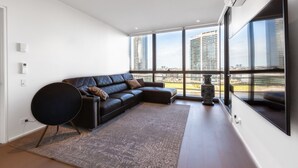 A modern living room with a sectional sofa, a large window with a city view, and a decorative vase. - Sabrina, 2BDR Docklands Apartment (Docklands)