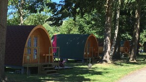 A row of wooden A-frame cabins with attached porches, set in a wooded area with a gravel path. - Camping Le Parc les Atypiques (Availles-Limouzine)