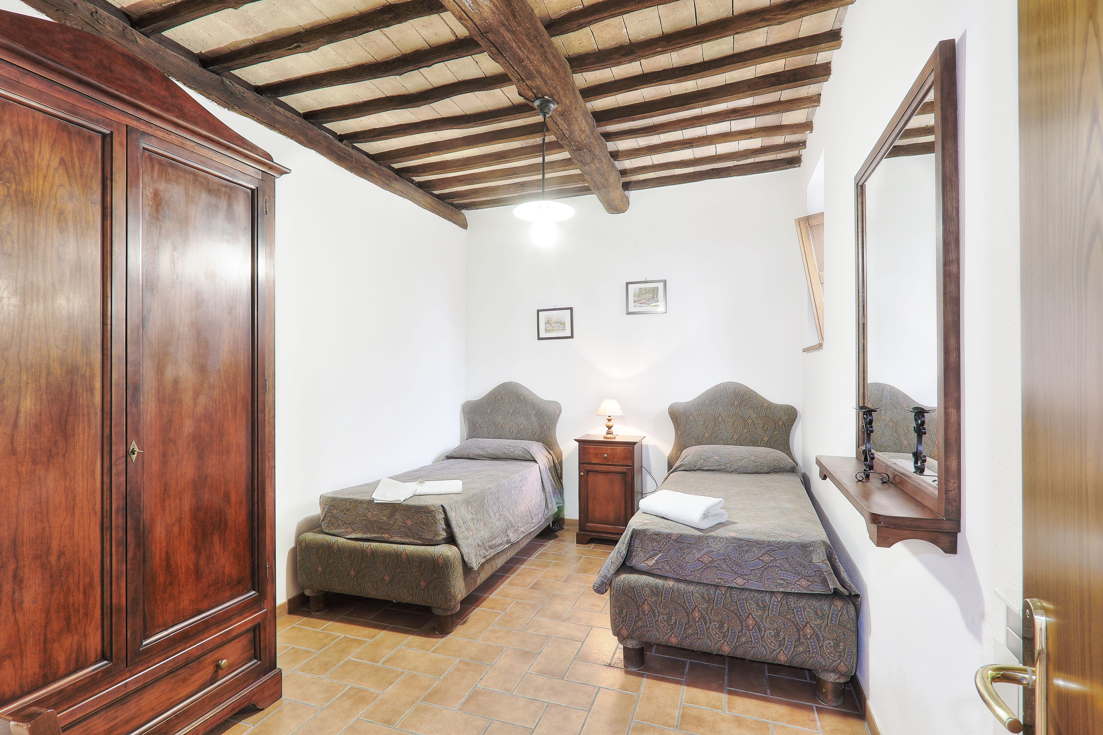 A bedroom with two single beds, a wooden wardrobe, a nightstand with a lamp, and a mirror on the wall.