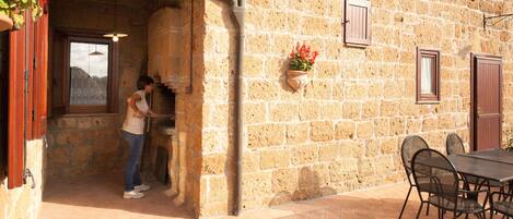 Un edificio de piedra con un área de estar al aire libre de estilo rústico, un horno de piedra y un ventanal con contraventanas.
