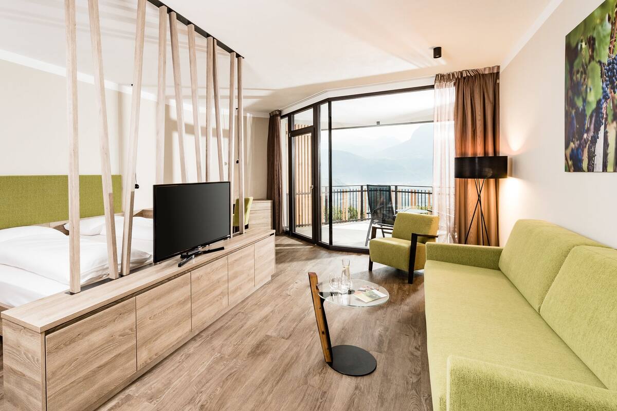A modern hotel room with a bed, TV, green sofa, and a balcony with a view.