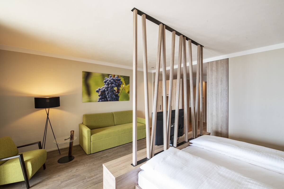 A hotel room with a bed, a green sofa, a floor lamp, and a painting of grapes on the wall.