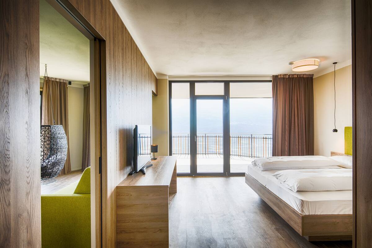 A modern hotel room with a large bed, a desk, a chair, and a view of the sea through large windows.