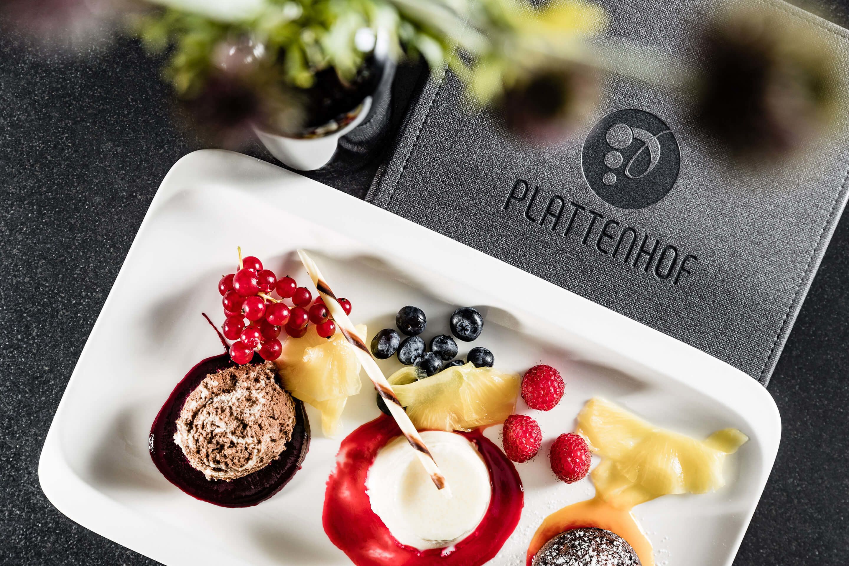 A white plate with various fruits and desserts, including red currants, blueberries, raspberries, and a slice of pineapple, on a dark surface.