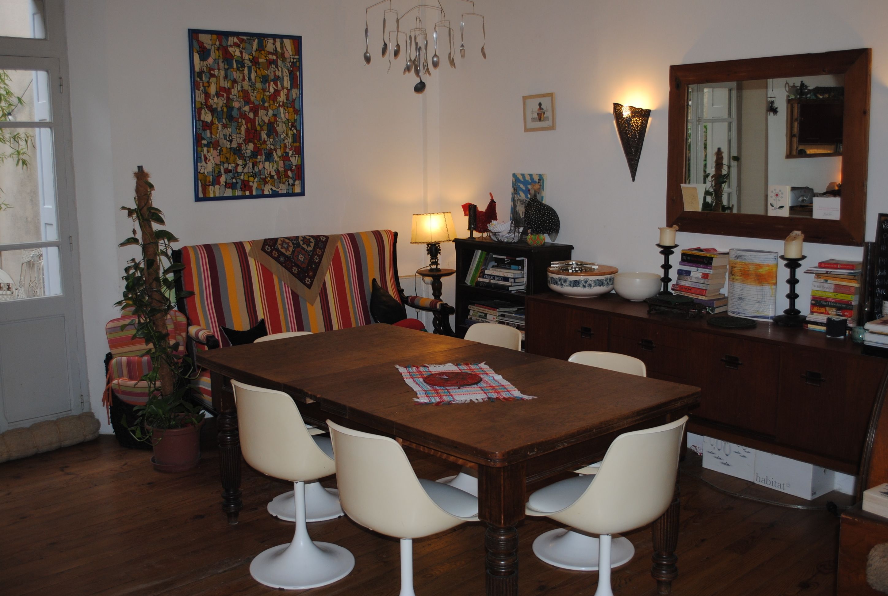 Un coin repas avec une table en bois, des chaises blanches, un canapé rayé aux couleurs vives, une plante en pot et une bibliothèque remplie de livres et d’objets décoratifs.
