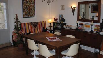 Un coin repas avec une table en bois, des chaises blanches, un canapé rayé aux couleurs vives, une plante en pot et une bibliothèque remplie de livres et d’objets décoratifs.